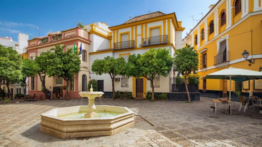 Calle estrecha y empedrada, flanqueada por fachadas blancas y balcones con flores en el Barrio de Santa Cruz, Sevilla.