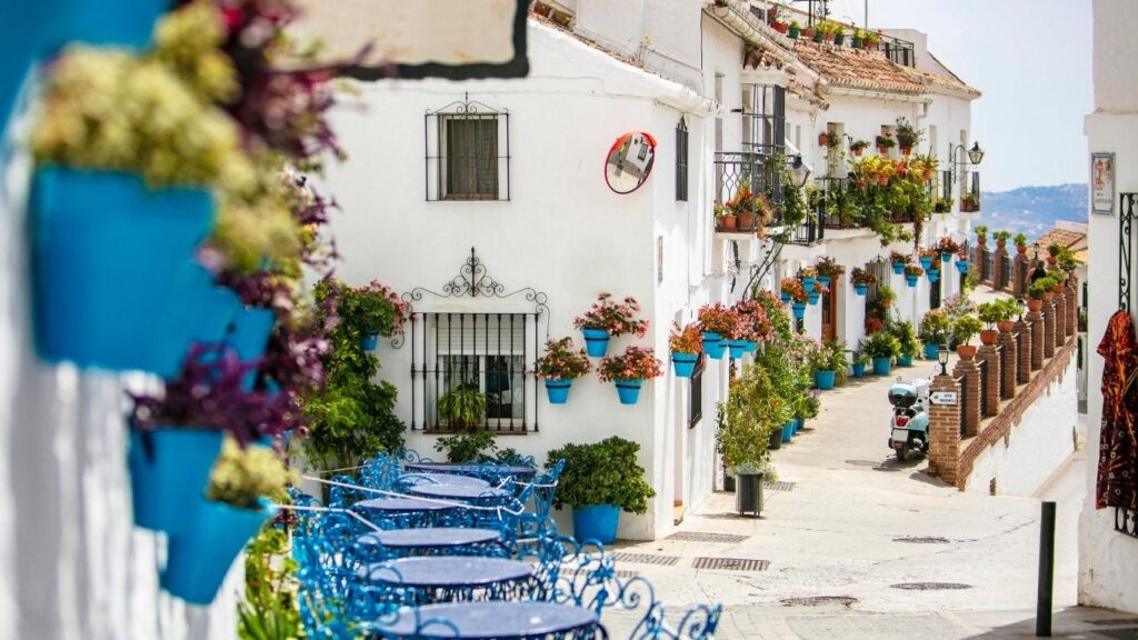 Calle empedrada de Mijas Pueblo con casas blancas decoradas con macetas de flores, mesas y sillas azules de terraza y un espejo convexo.