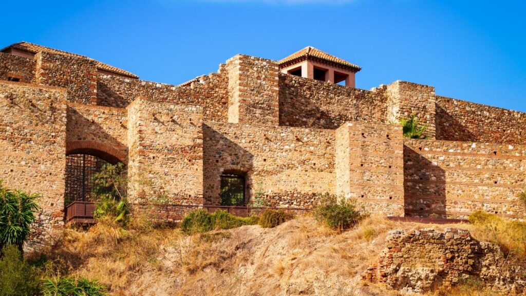 Murallas de piedra rojiza del Castillo de Gibralfaro en Málaga, con un torreón y almenas, bajo un cielo azul despejado.