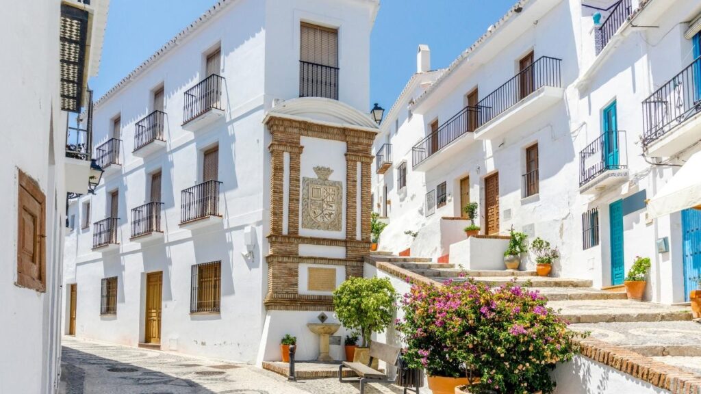 Calle estrecha y empinada del pueblo blanco de Frigiliana, con casas encaladas, macetas con flores y escaleras de piedra.