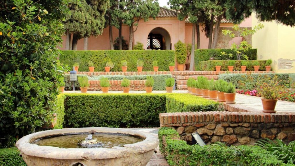 ardines de la Alcazaba de Málaga con setos verdes cuidadosamente podados, fuentes de piedra y una estructura árabe al fondo.