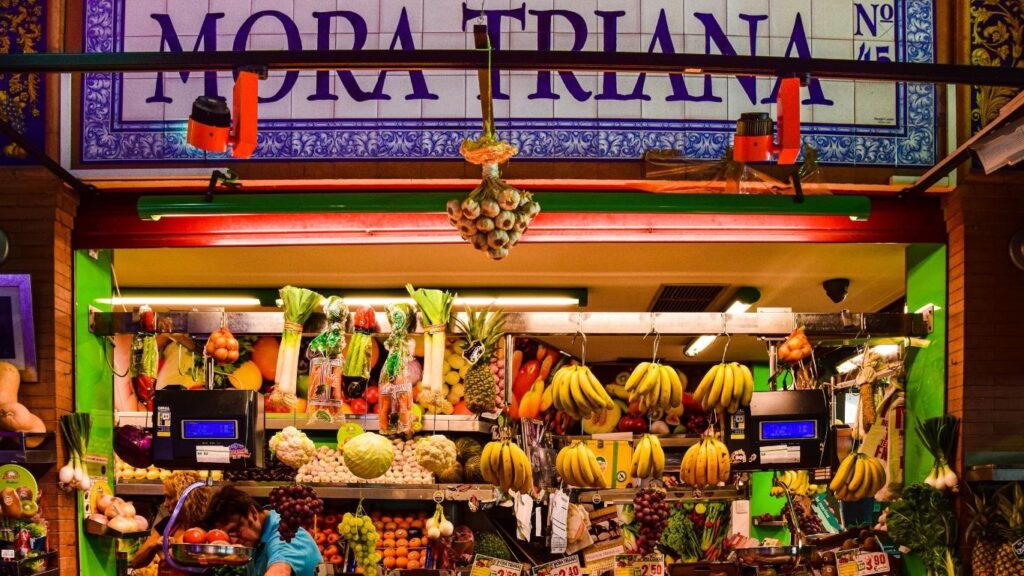 Detalle de un puesto de frutas y verduras frescas en el Mercado de Triana, con el letrero 'Mora Triana' en azulejos.