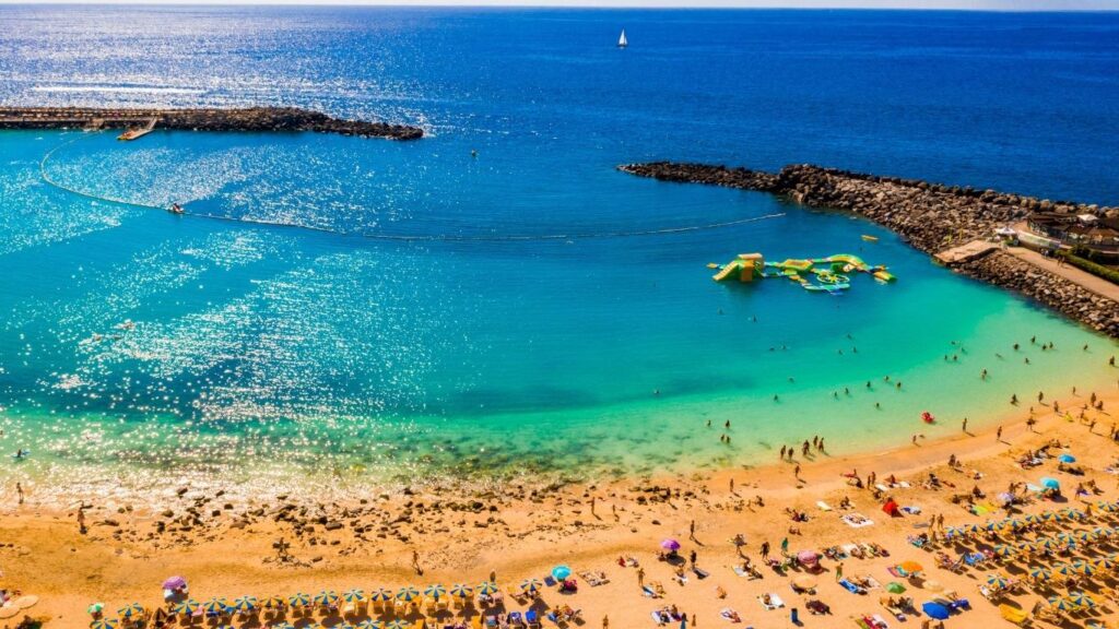 Vista panorámica de la Playa Amadores en Gran Canaria, destacando sus aguas turquesas y los espigones artificiales que la protegen del oleaje.