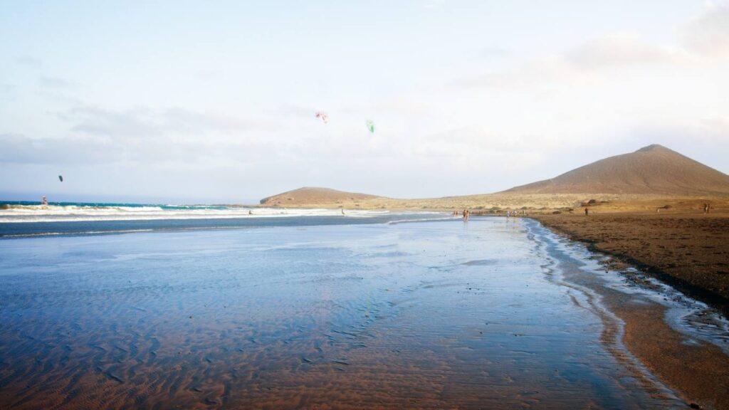 Playa El Médano en Tenerife con viento, mostrando la marea baja y aficionados al windsurf y kitesurf en la distancia.