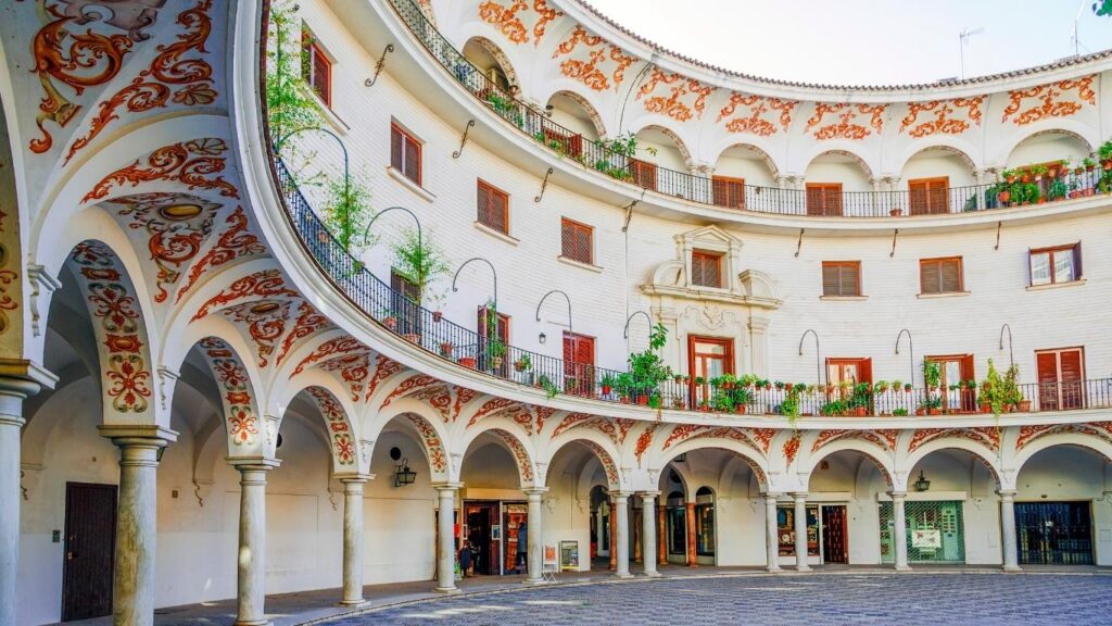Arquitectura semicircular de la Plaza del Cabildo en Sevilla, con arcos blancos y detalles decorativos en naranja, vista desde el patio.