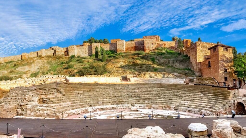 Gradas semicirculares del Teatro Romano de Málaga en primer plano, con las murallas de la Alcazaba elevándose en la colina de fondo.