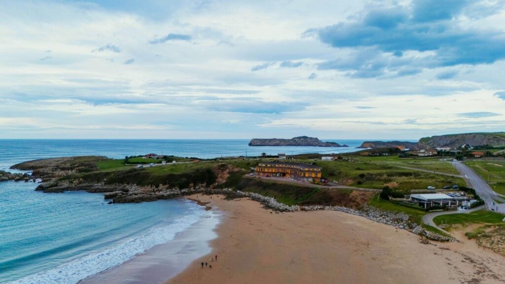 Vista panorámica de la Playa de Oyambre con prados verdes, dunas y el mar bajo un cielo nublado en Cantabria.