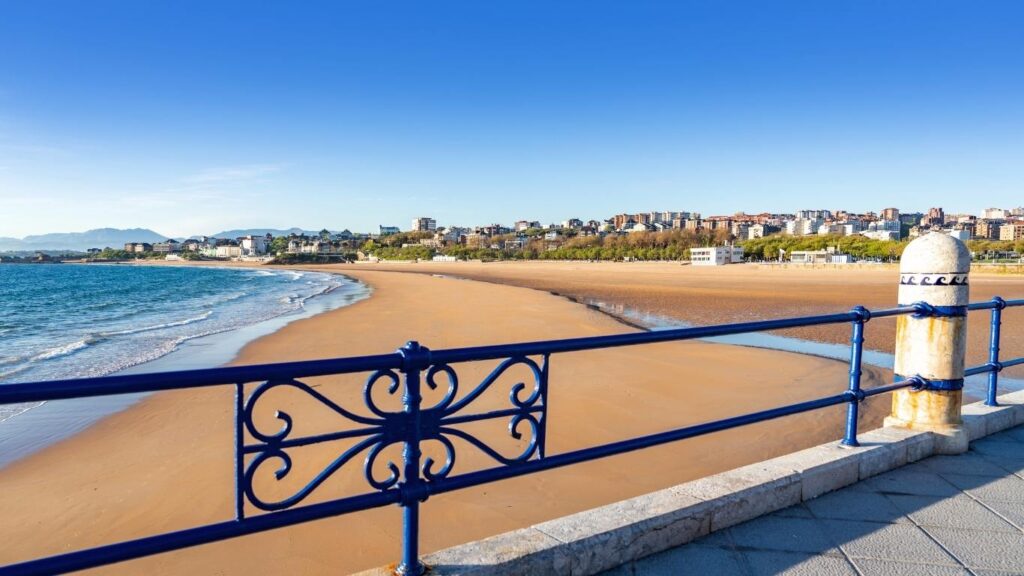 Valla azul de hierro en primer plano con vistas a la arena dorada de la Playa del Sardinero en Santander bajo un cielo despejado.