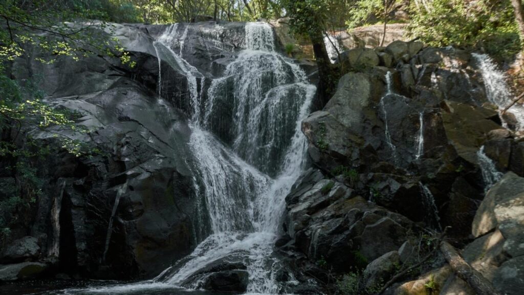 Un salto de agua natural que se desliza por una pared de roca rojiza con musgo verde, rodeada de vegetación densa y silvestre en Navaconcejo.