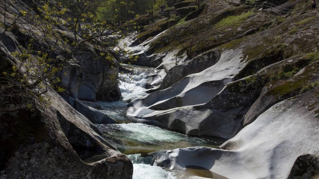 El río Jerte fluyendo entre grandes formaciones de granito liso y redondeado que forman pozas naturales (pilones), con vegetación de ribera y senderos en las laderas.