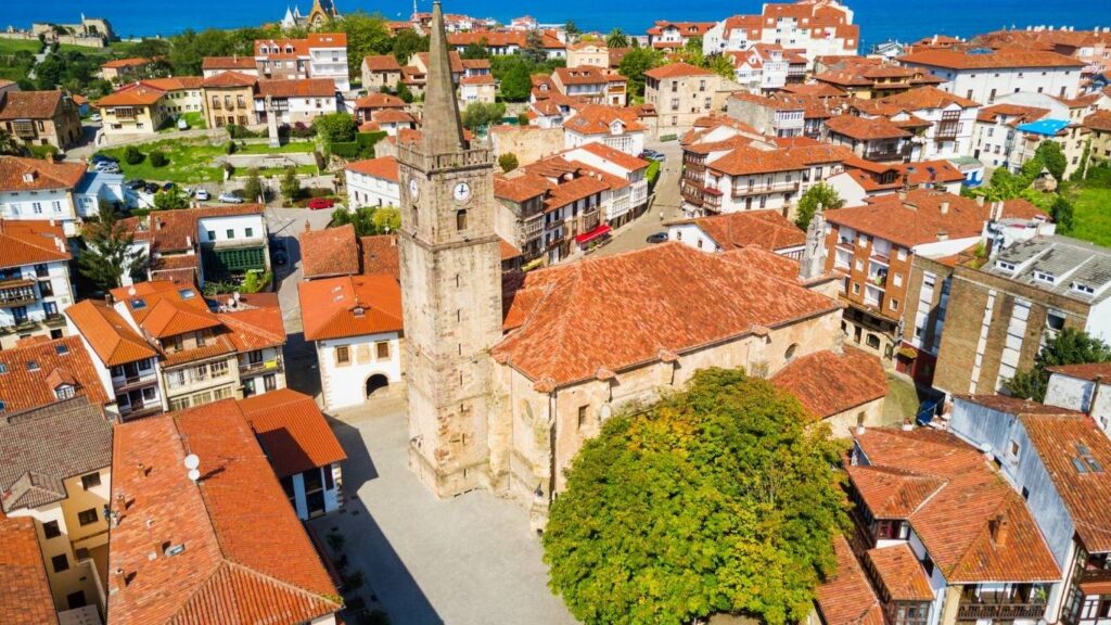 Vista aérea de la Iglesia de San Cristóbal en Comillas destacando su torre y los tejados rojos del pueblo.