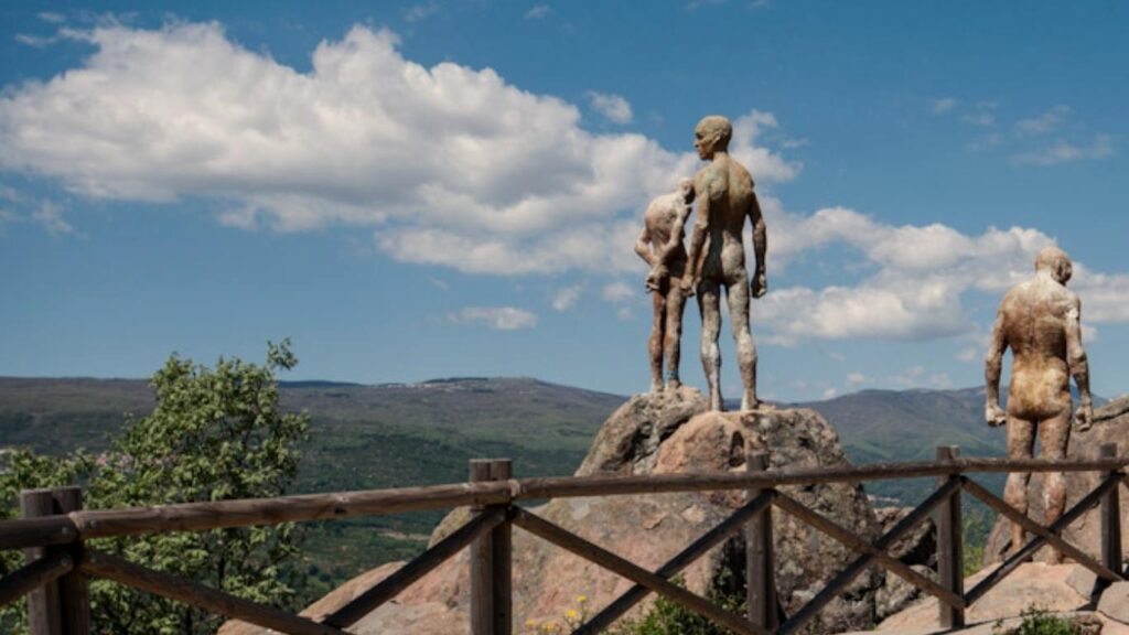 Escultura de figuras humanas desnudas de estilo contemporáneo sobre una roca, con el valle extremeño y montañas al fondo desde un mirador de madera.