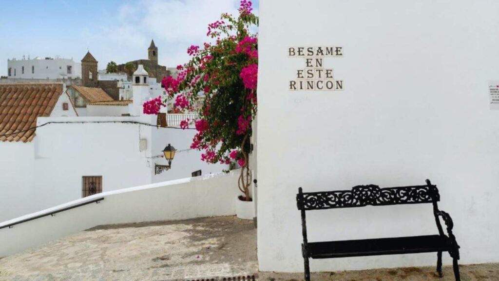 Un banco de forja junto a una buganvilla rosa y un cartel en la pared que dice "Bésame en este rincón" en Vejer.